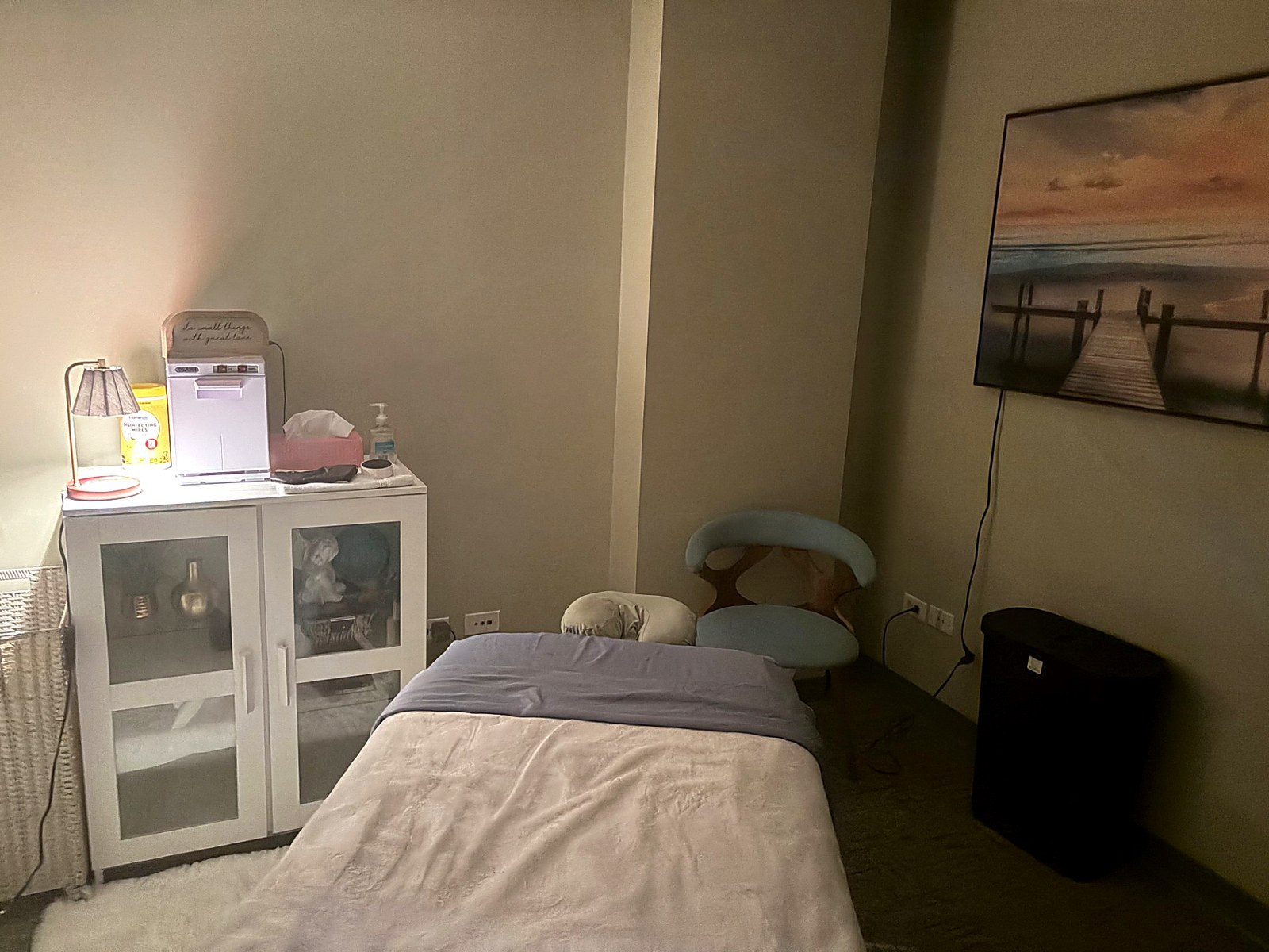 The private room with a single massage table made up in soft linens, a white cabinet, warm lamp light and a framed pier photograph on the wall